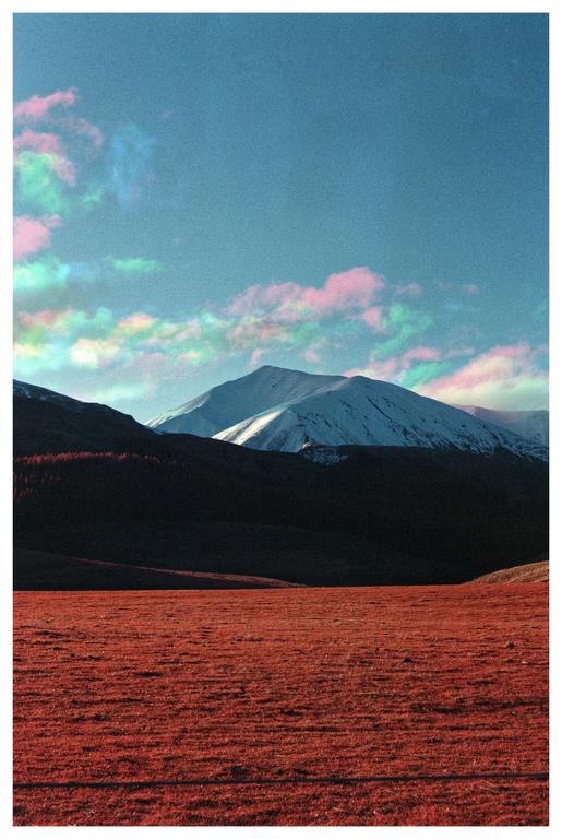 A red field sits in front of dark hills and a snow-capped mountain under a blue sky with colorful clouds.