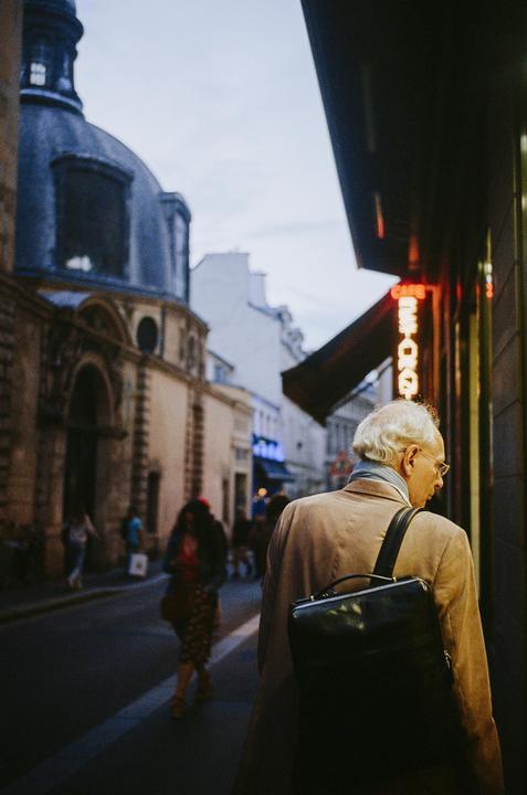An older man with white hair and glasses walks away from the viewer on a street with buildings and other people in the background.