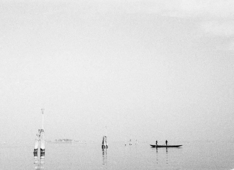 Two people stand in a small boat on the water with several wooden posts visible.