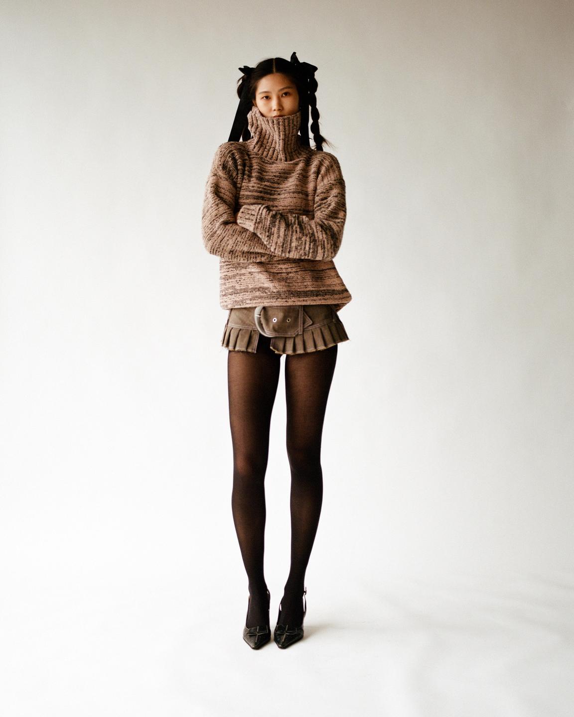 A woman with braided pigtails and a turtleneck sweater stands with her arms crossed.
