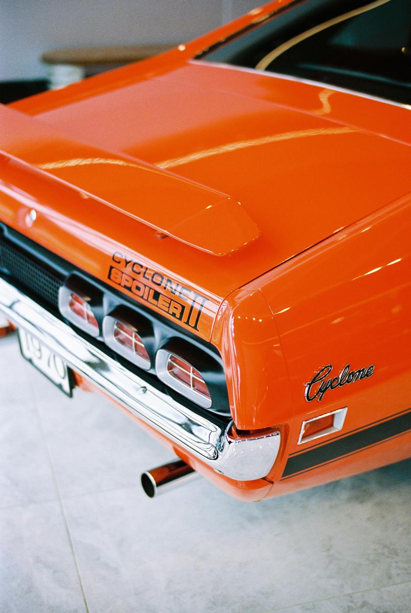 The rear of an orange Mercury Cyclone car is visible, showing the taillights, bumper, and exhaust pipe.