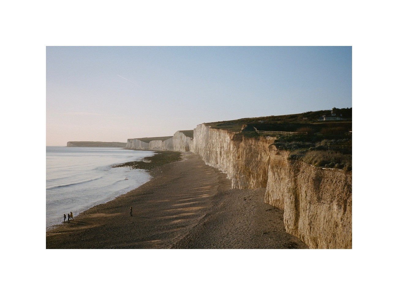 A long stretch of white cliffs rises above a beach and the sea, with a few people visible on the shore.