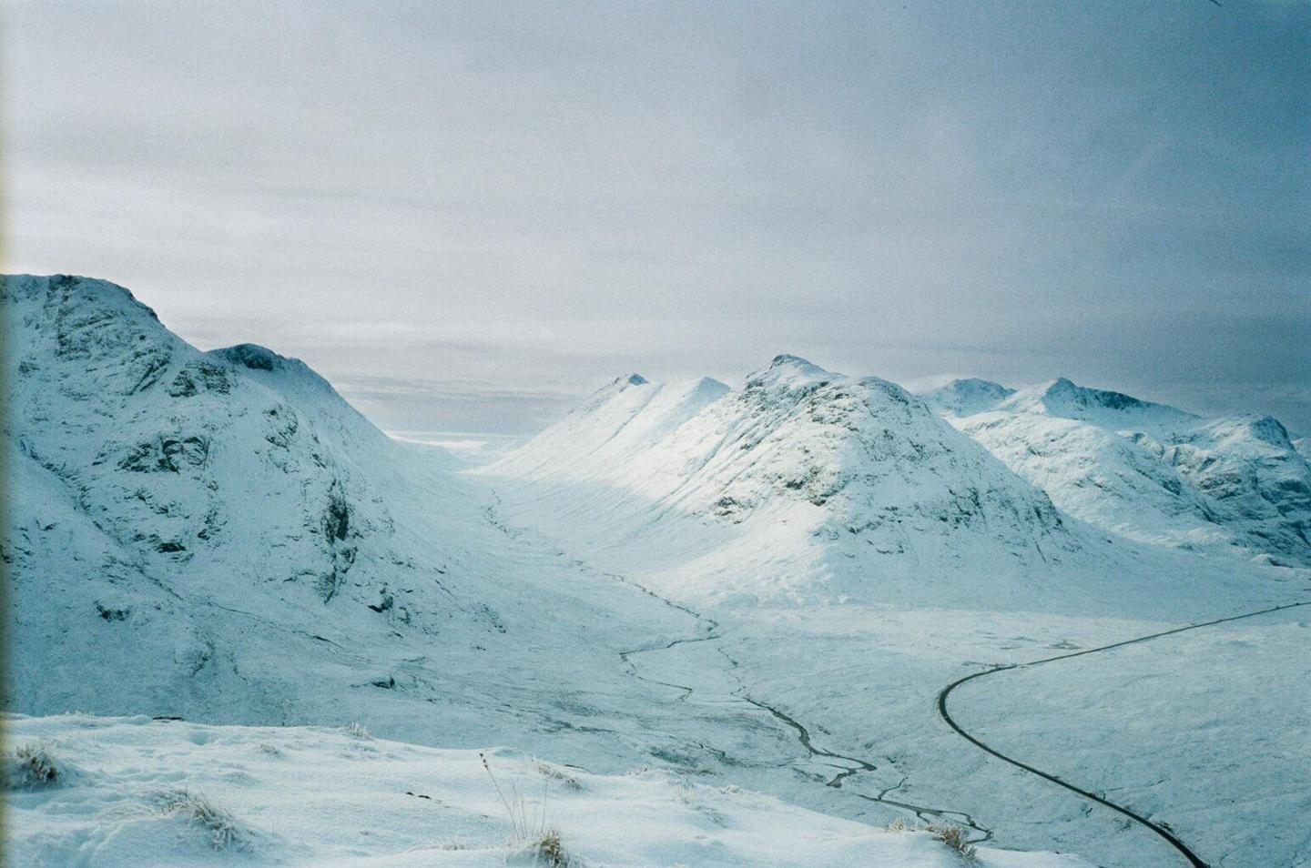 A winding road cuts through a snow-covered valley surrounded by mountains under a cloudy sky.