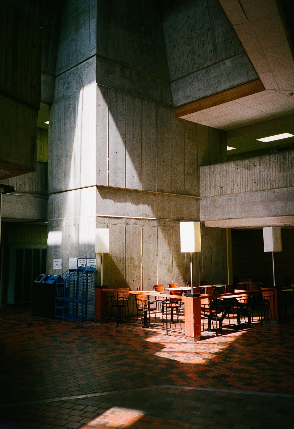 Sunlight streams into a brutalist building with concrete walls, a tiled floor, and a seating area with tables and chairs.