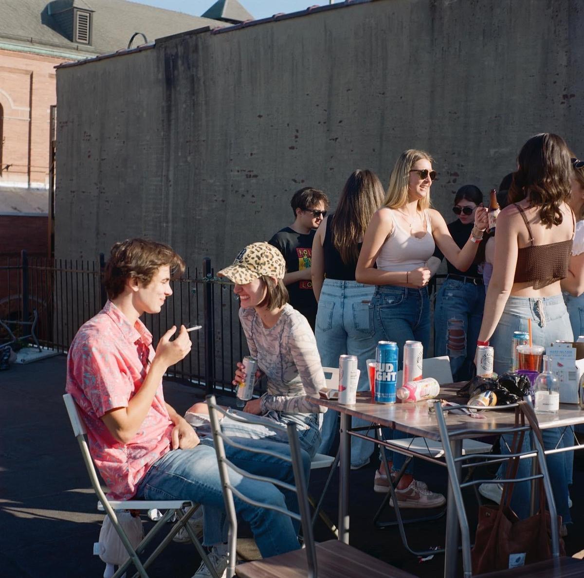 A group of young adults are gathered around a table with drinks, with one man smoking a cigarette while talking to a woman in a leopard print hat.