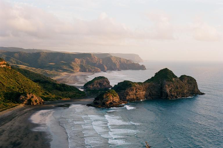A wide view shows a beach and ocean with waves next to a series of green hills and rocky cliffs.