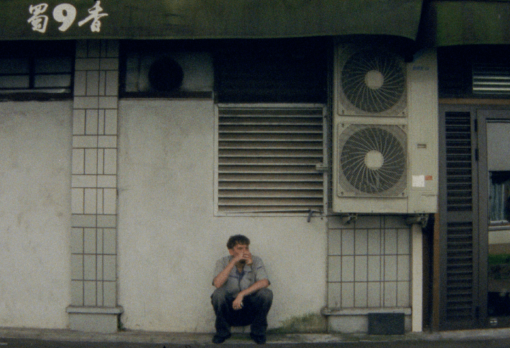 A man crouches on a sidewalk next to a building with air conditioning units and a sign with Asian characters.