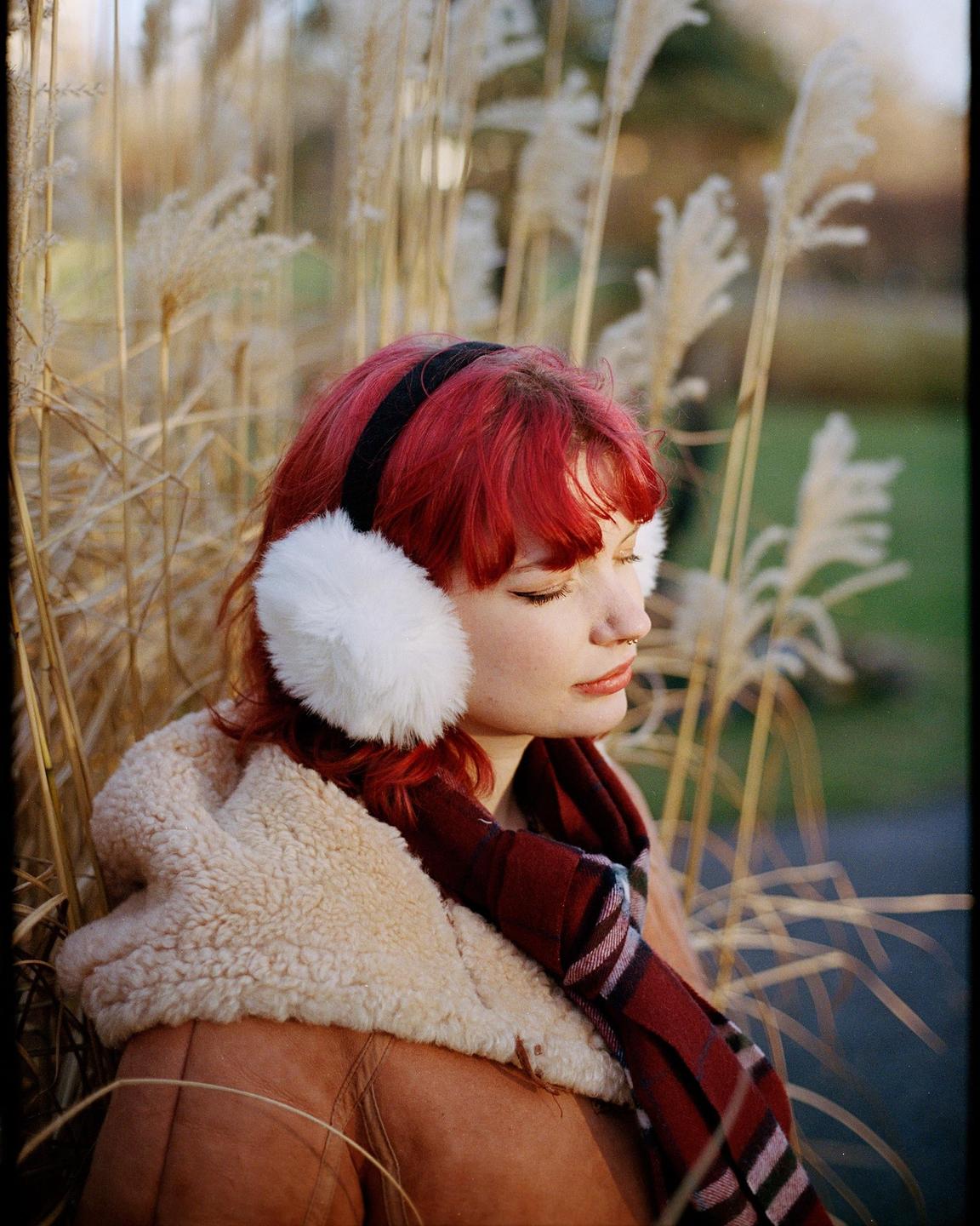 A woman with red hair and white earmuffs is sitting among tall grasses.