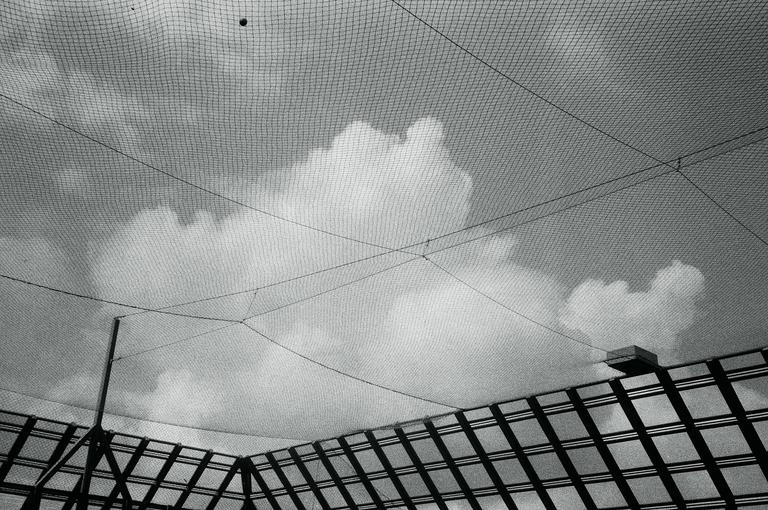 A baseball net covers the sky with clouds and a building visible below.