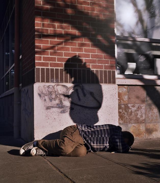 A person is lying on the sidewalk next to a brick building with graffiti and a large shadow of a person on the wall.