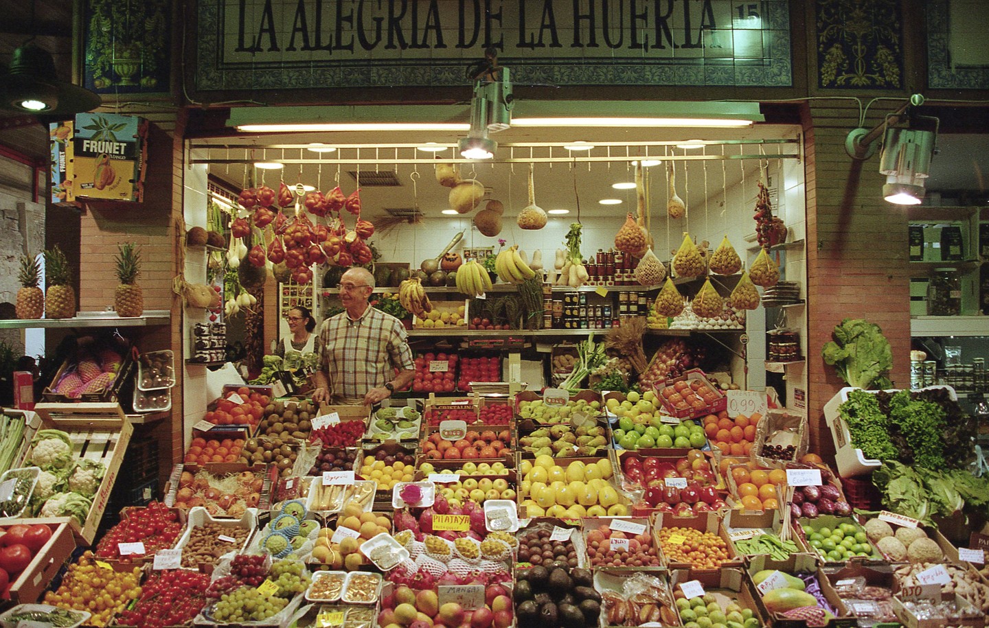 A man stands behind a vibrant fruit and vegetable stall with a woman in the background, under a sign that reads "LA ALEGRIA DE LA HUERTA."