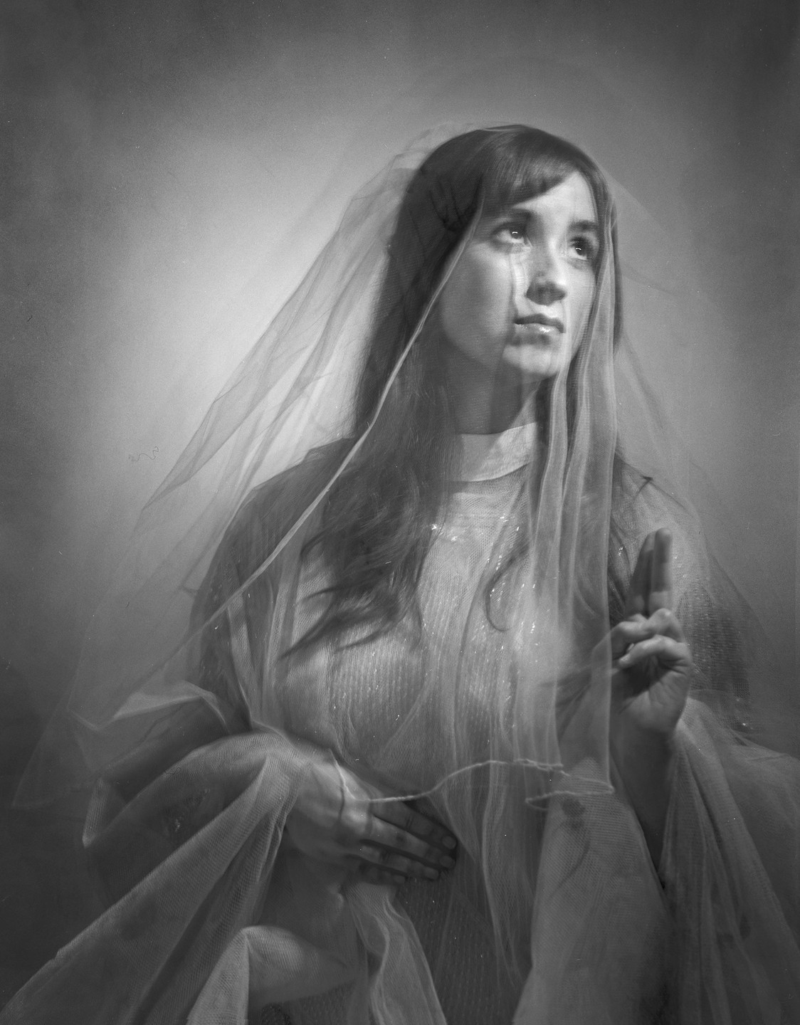 A woman with long hair and bangs wears a sheer veil and a textured dress, looking upwards with a tear on her cheek and one hand raised in a gesture.