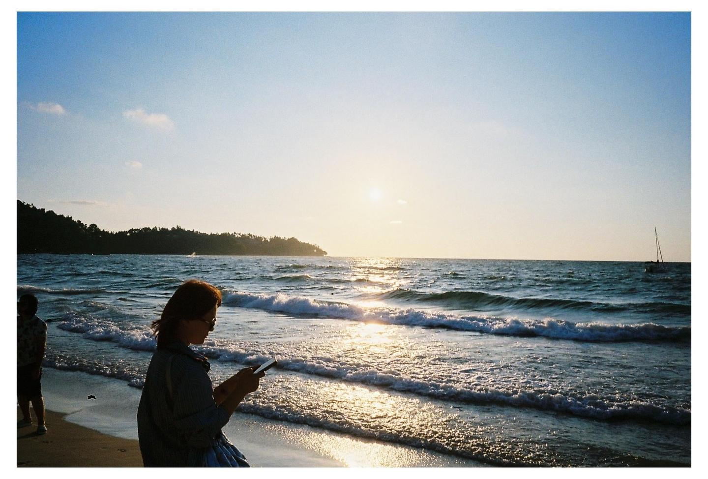 A woman sits on a beach looking at her phone with the ocean and a distant sailboat in the background.