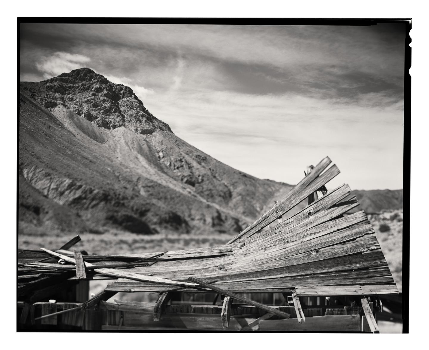 A dilapidated wooden structure sits in the foreground with a large mountain in the background under a cloudy sky.