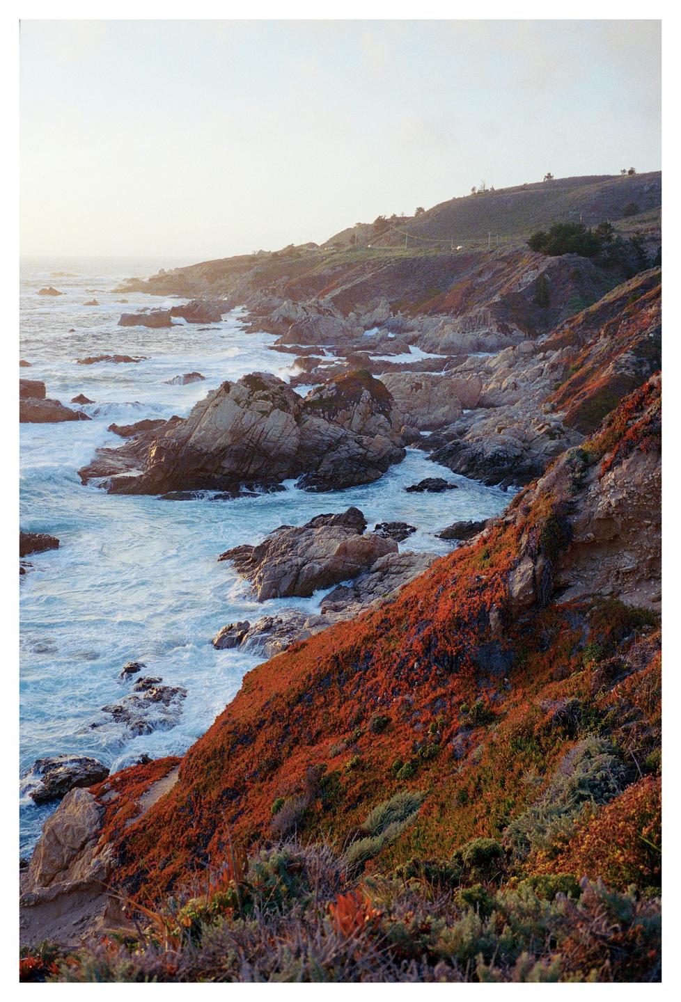 Waves crash against a rocky coastline with hills covered in red and green vegetation under a bright sky.