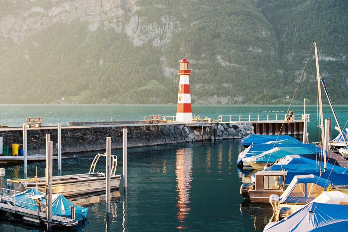 A red and white striped lighthouse stands on a pier with boats docked in the foreground and a mountain in the background.