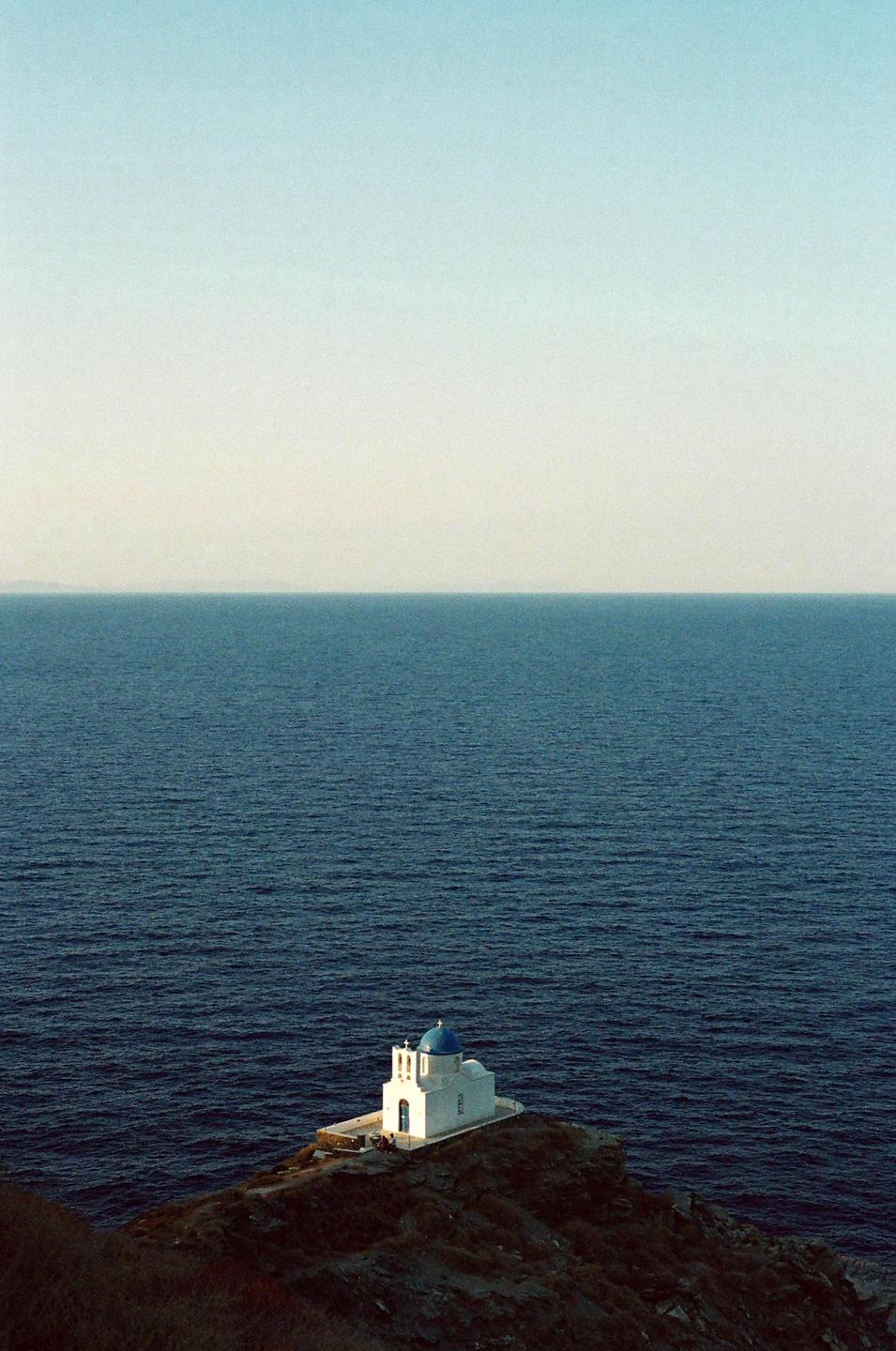 A white church with a blue dome sits on a cliff overlooking the ocean.