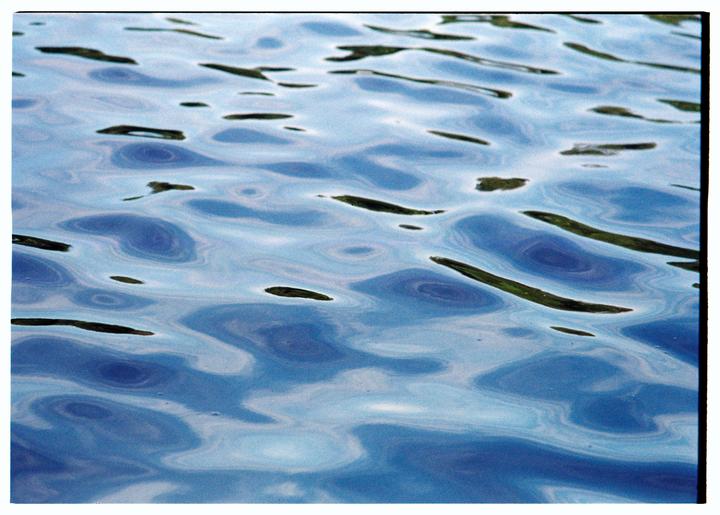 The surface of a body of water shows ripples and reflections of blue and green.