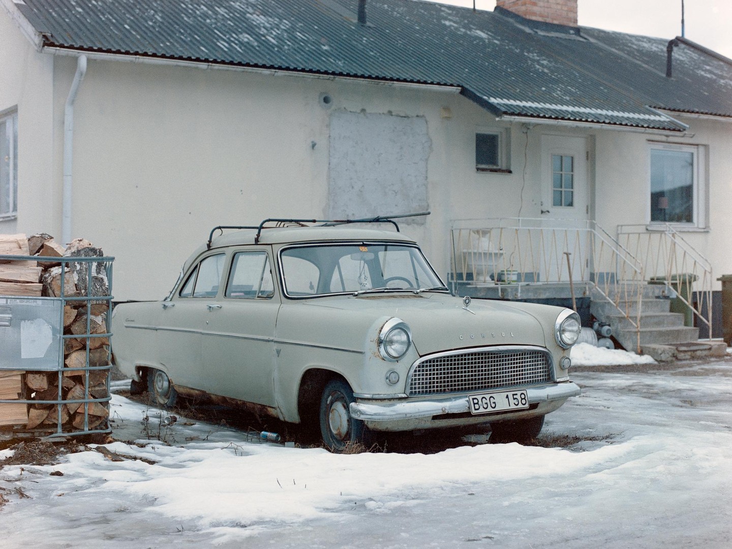 A light-colored vintage car with a roof rack is parked in front of a house with snow and ice on the ground.