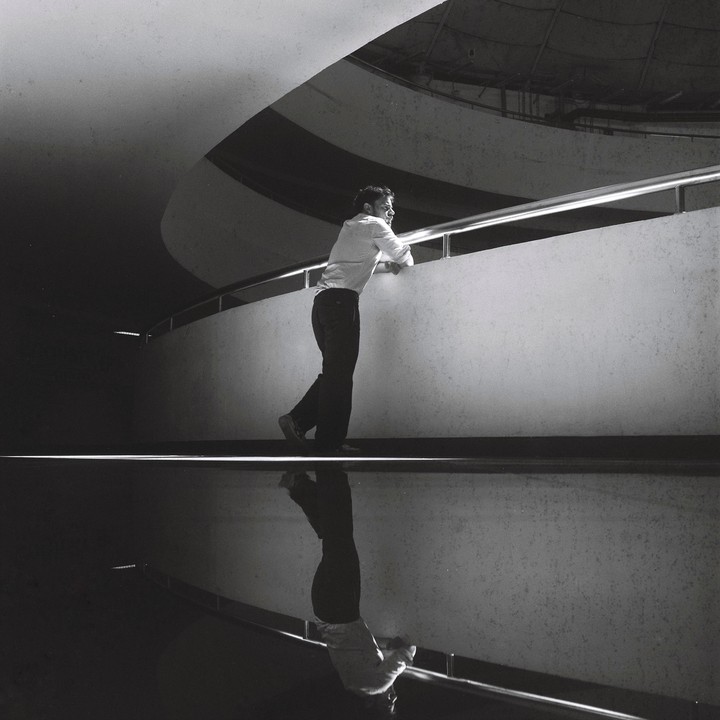 A man leans on a railing with his reflection visible below him.