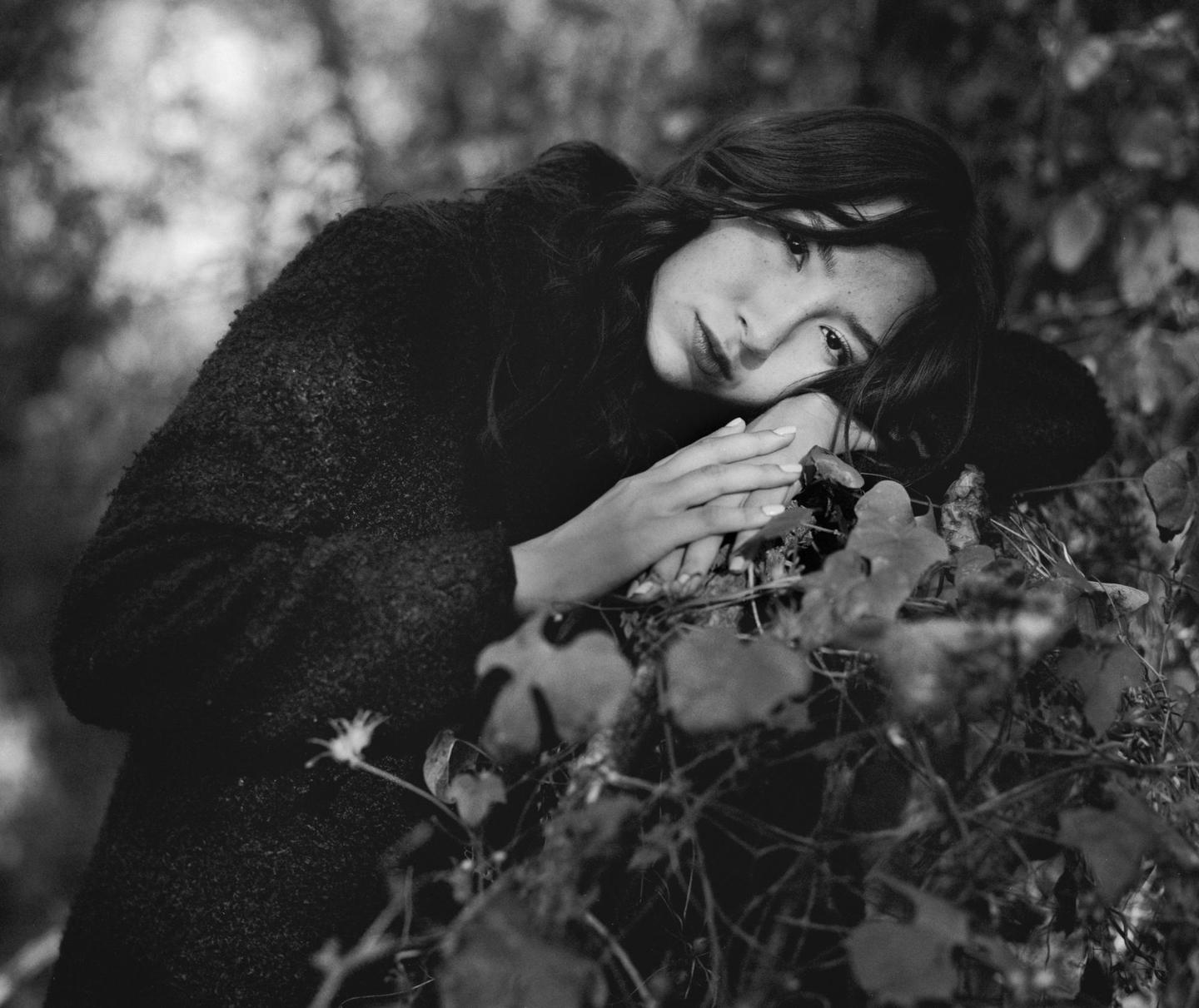 A woman with dark hair is resting her head on her hands among foliage.