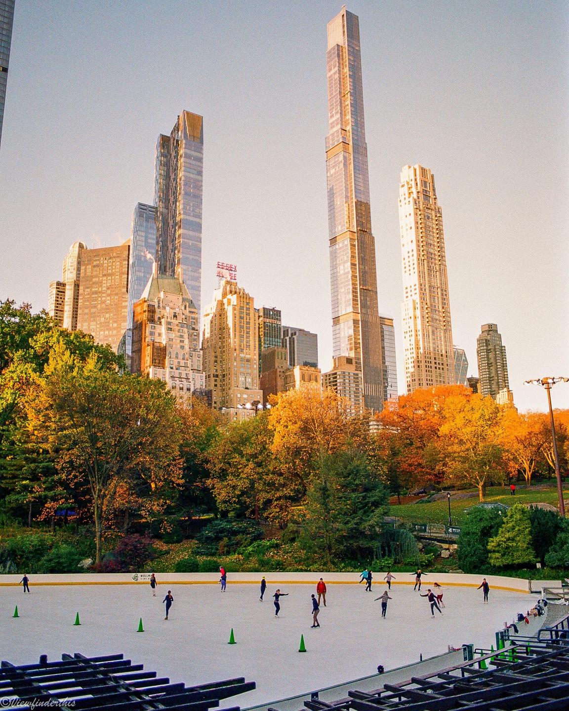People are ice skating on a rink surrounded by autumn trees and city buildings.