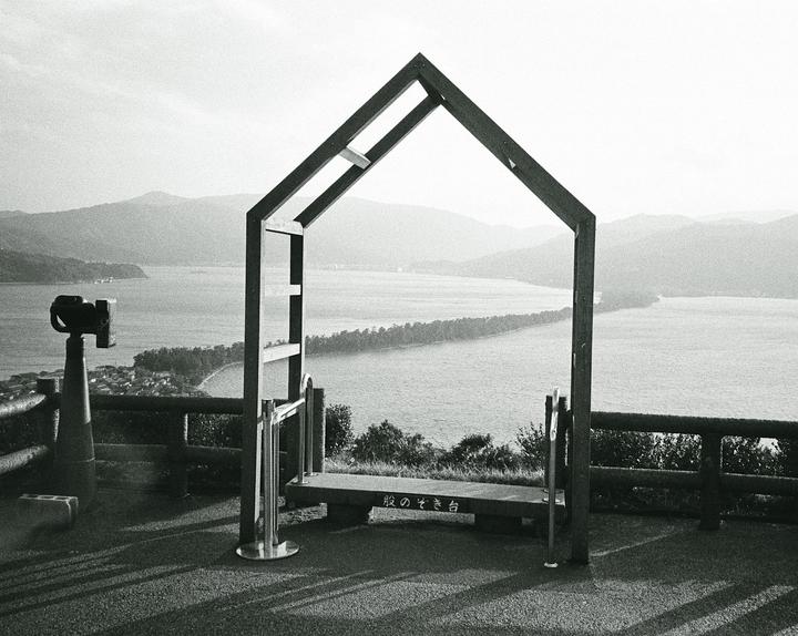 A wooden frame shaped like a house stands on a hill overlooking a body of water with a narrow land bridge and mountains in the distance.