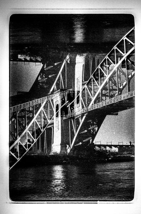 A black and white photograph captures the intricate structure of a bridge spanning over water, with reflections visible on the surface.