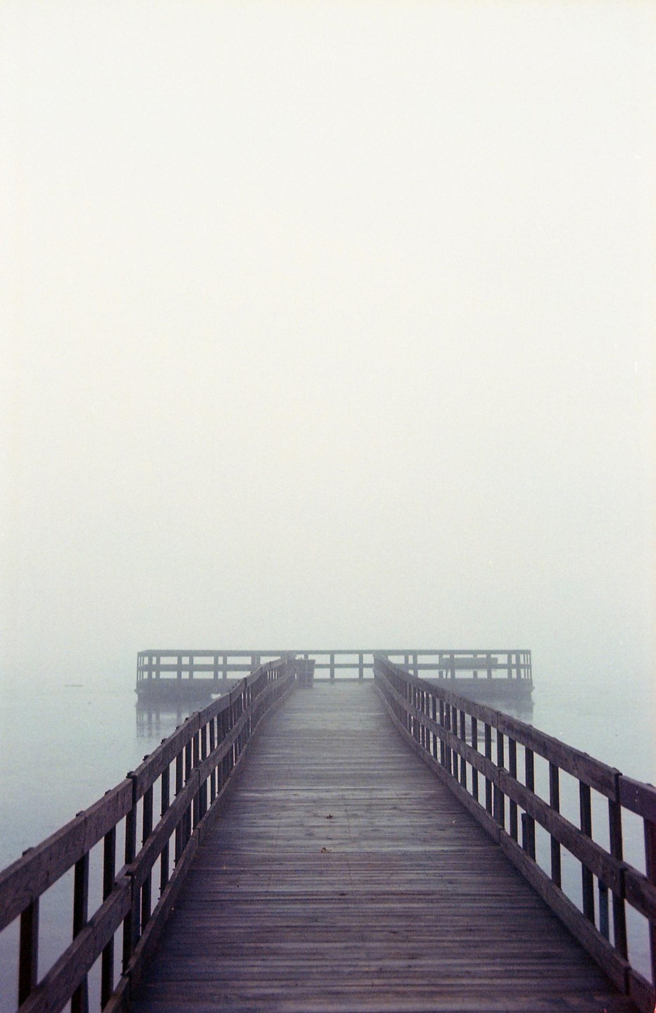 A wooden pier extends into a foggy lake, leading to a circular platform at its end.