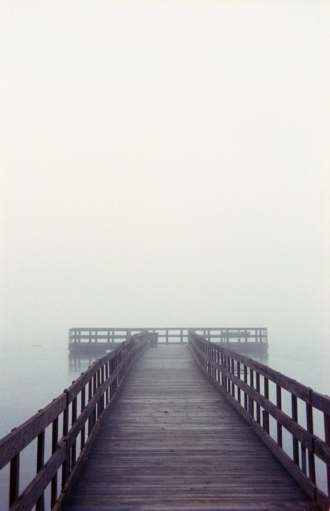 A wooden pier extends into a foggy lake, leading to a circular platform at its end.