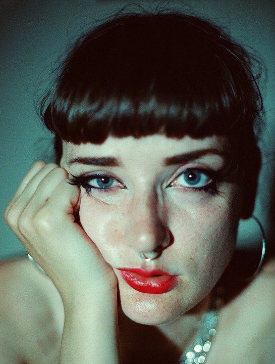 A woman with bangs, red lipstick, and a nose ring looks directly at the viewer.