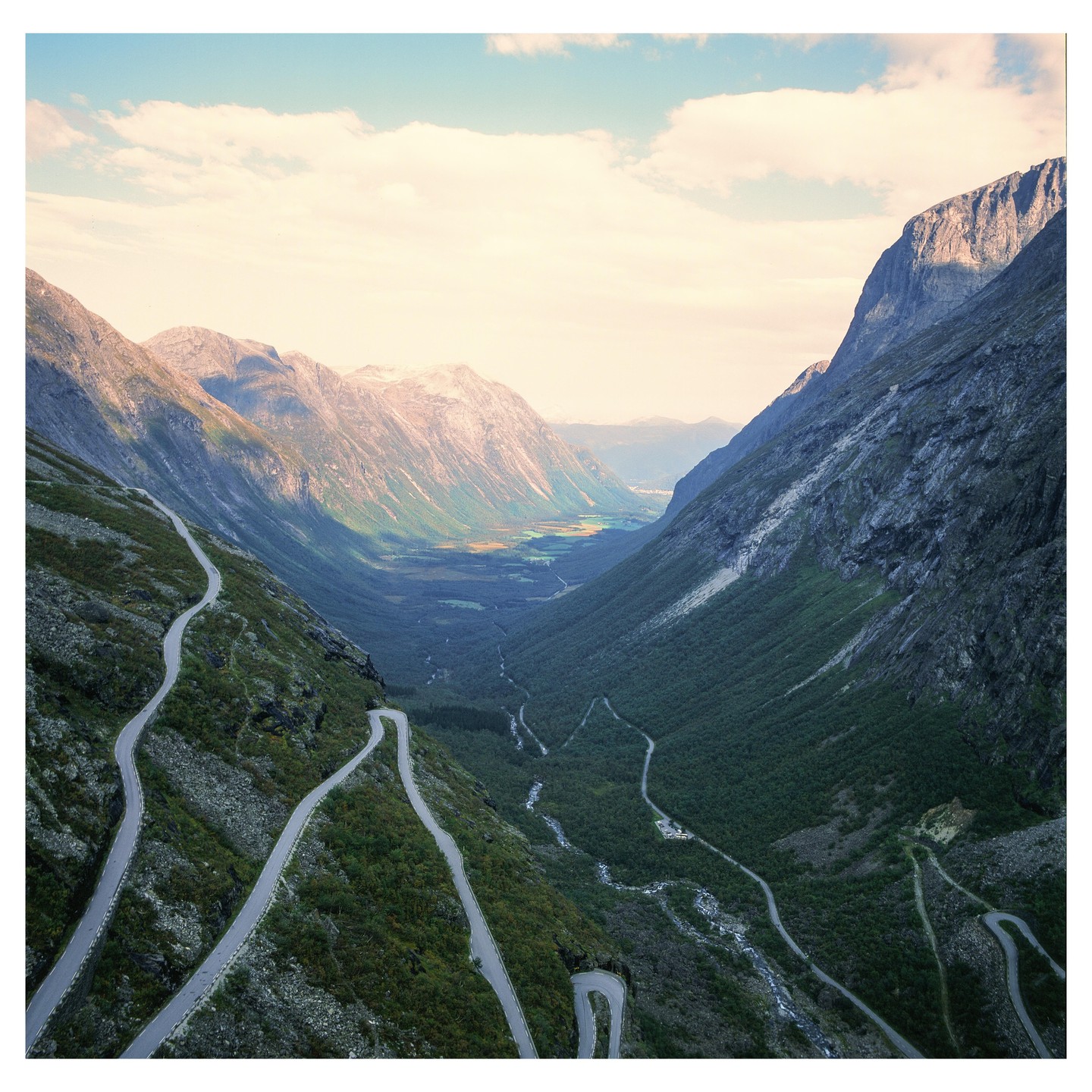 A winding road descends into a valley surrounded by mountains under a cloudy sky.