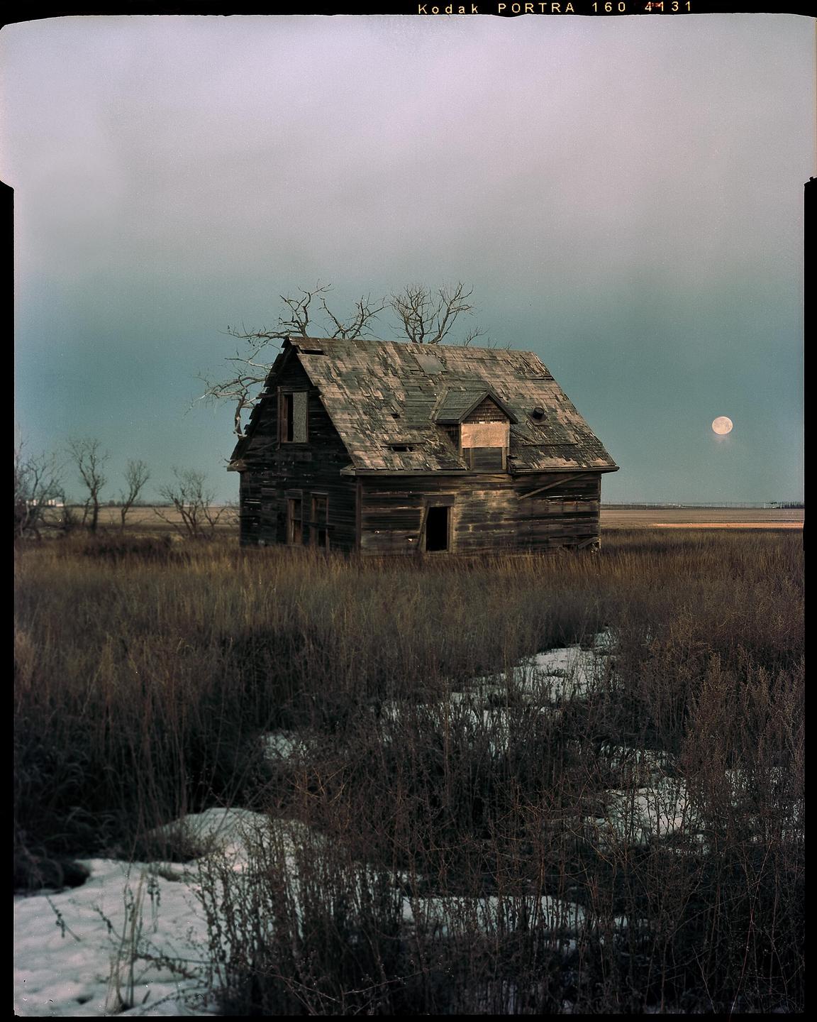 An abandoned wooden house stands in a field with patches of snow under a cloudy sky with a full moon.