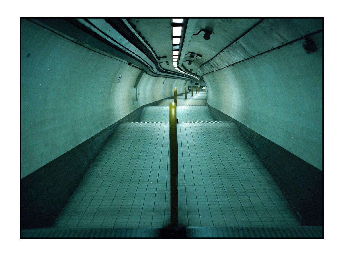A long, empty underground tunnel with a tiled floor and yellow poles leading into the distance.