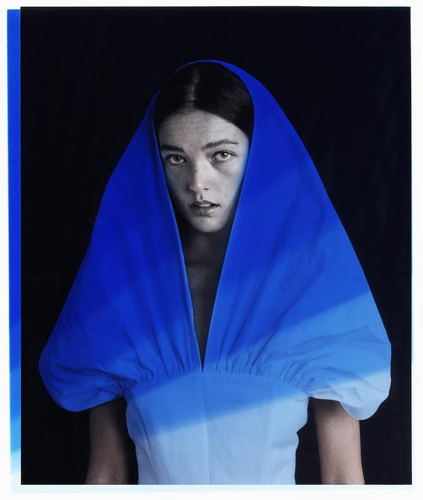 A woman with dark hair and freckles wears a blue and white garment with a hood.