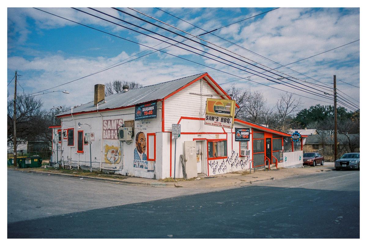 A white building with red trim and a sign for Sam's BBQ stands on a street corner under a blue sky with power lines overhead.
