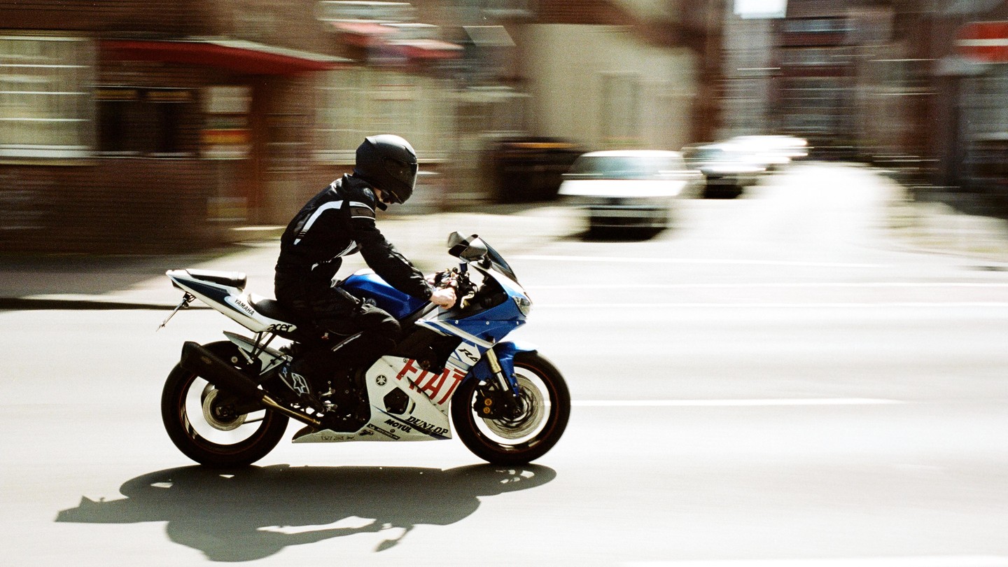 A person on a blue and white motorcycle speeds down a street, with buildings and cars blurred in the background.