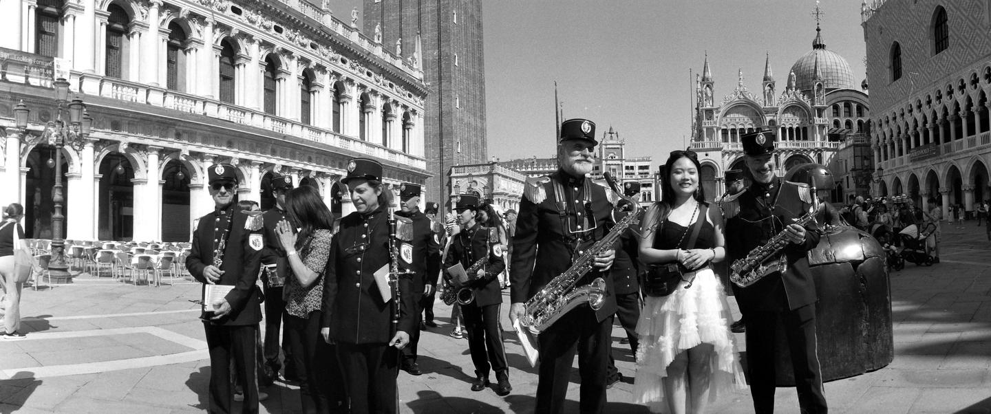 A group of musicians in uniform and a woman in a white dress stand in a square in front of ornate buildings.