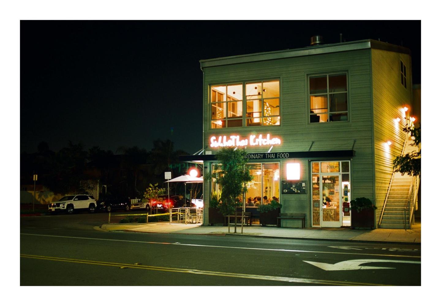 A brightly lit Thai restaurant with a neon sign stands on a street corner at night.
