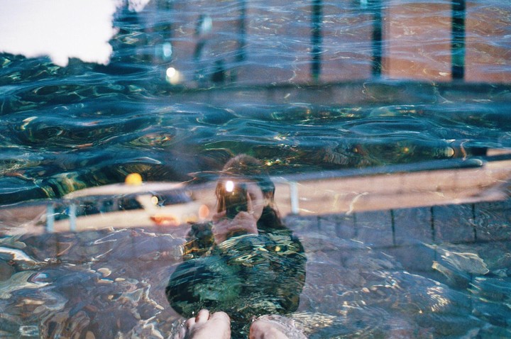 A self-portrait is visible through the rippling surface of water, with feet submerged in the foreground.