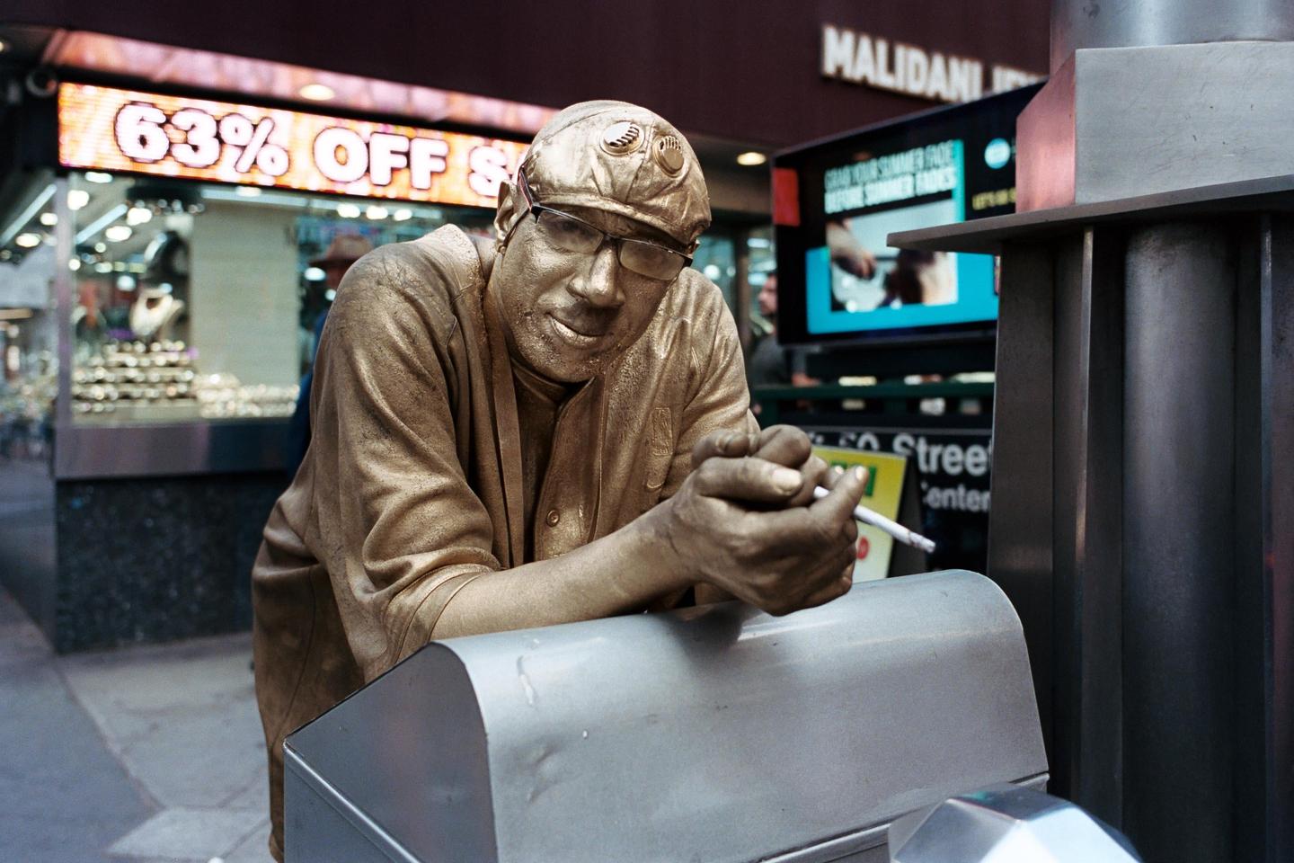 A man covered in gold paint leans on a metal box on a city street.