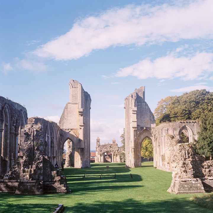 The ruins of an abbey stand on a green lawn under a blue sky with white clouds.