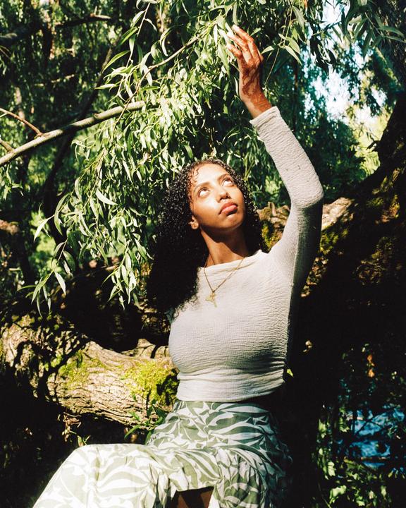 A woman with dark curly hair and a white top sits on a mossy tree branch, reaching up towards green leaves.