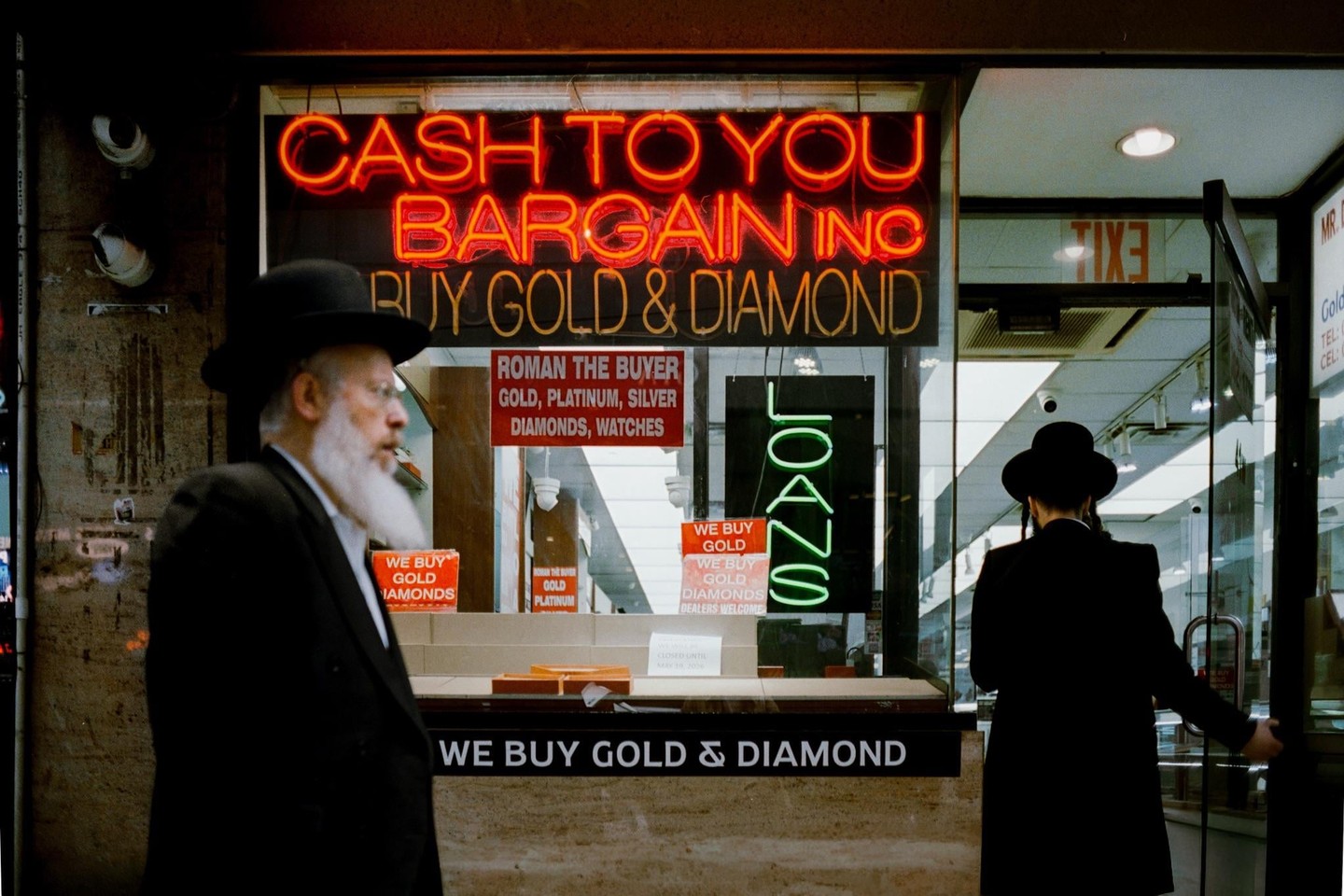 Two men in black hats and coats stand in front of a shop with neon signs that read "Cash to You Bargain Inc" and "Buy Gold & Diamond."