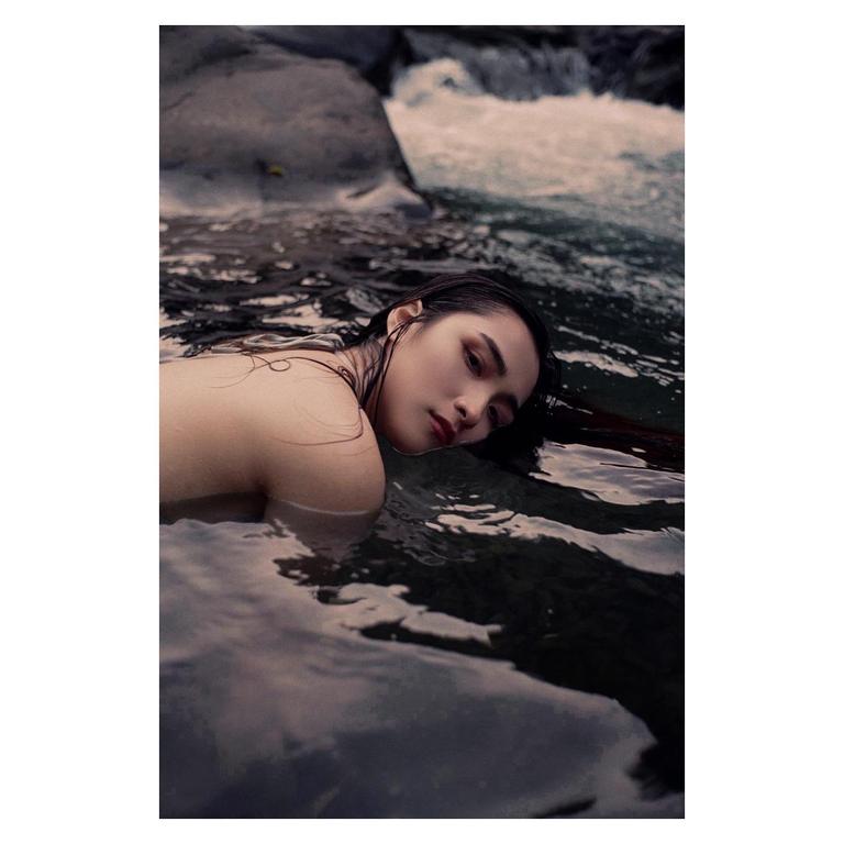 A woman with wet hair is partially submerged in water, looking towards the viewer.