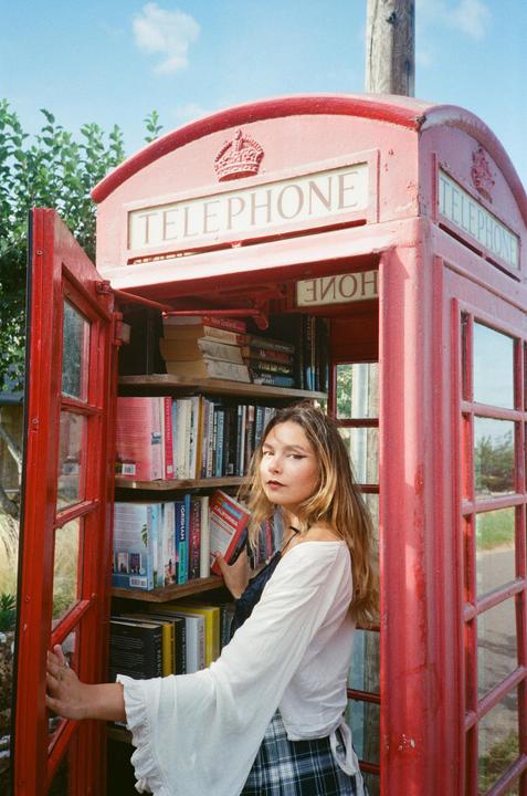 A woman stands inside a red telephone booth that has been converted into a community library, holding a book.