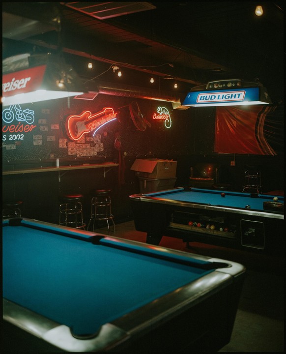 Two pool tables are visible in a dimly lit room with neon signs on the wall.