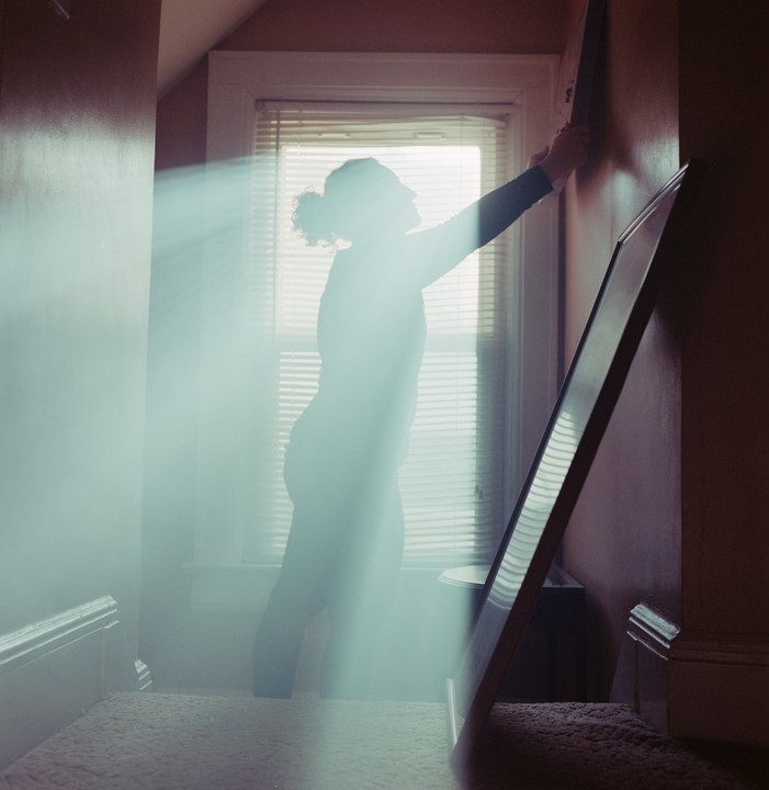 A woman is silhouetted by light from a window as she hangs a picture frame on a wall.