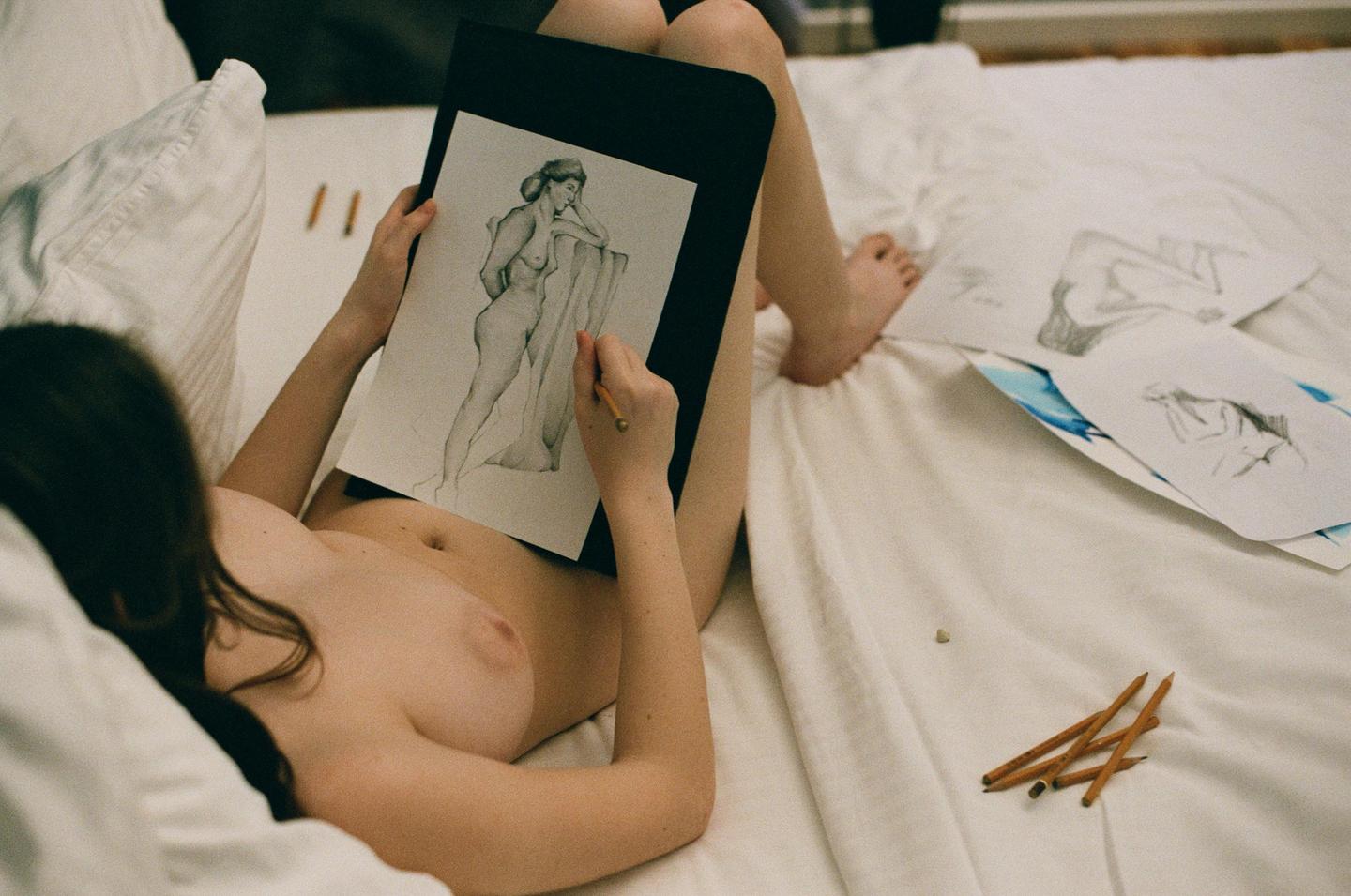 A woman lies on a bed drawing a nude figure with a pencil.