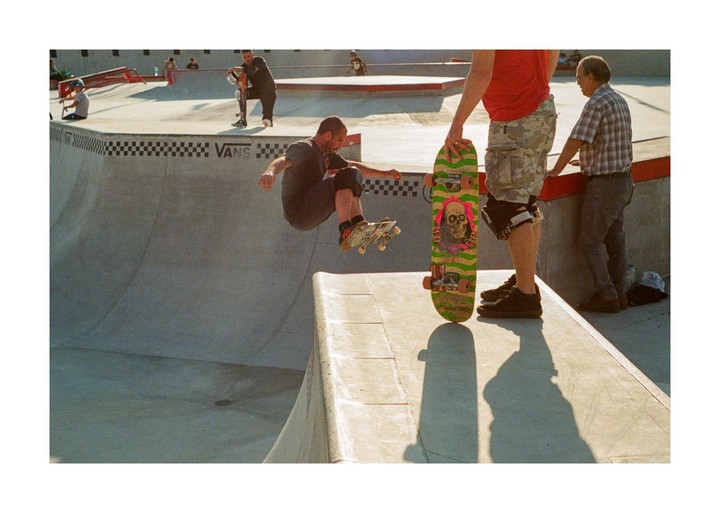 A man on a skateboard jumps in a skatepark while another man stands nearby with a skateboard.